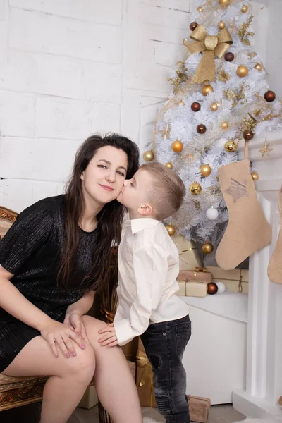 Atmosphere of coming holidays. portrait of happy single mother with her little pleasant boy in white bright room near christmas tree in golden tones — Stock Fotó