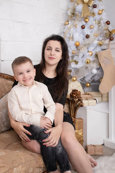 Atmosphere of coming holidays. portrait of happy single mother with her little pleasant boy in white bright room near christmas tree in golden tones — Stockfoto