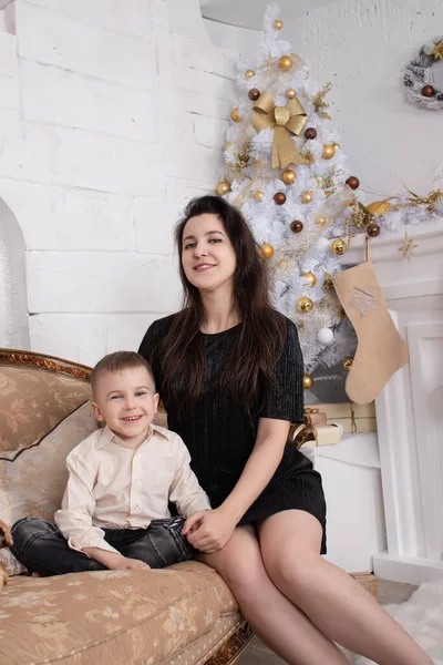 Atmosfera das próximas férias. retrato de mãe solteira feliz com seu menino agradável no quarto brilhante branco perto da árvore de natal em tons dourados — Fotografia de Stock