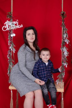 Mutlu eksik aile kucaklaşması ahşap salıncakta kırmızı arka planda noel ağacı dallarıyla süslenmiş. Sıcak, genç bir aile. Gri elbiseli bekar anne ve küçük oğlu.