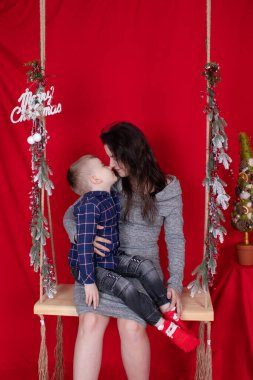 Mutlu eksik aile kucaklaşması ahşap salıncakta kırmızı arka planda noel ağacı dallarıyla süslenmiş. Sıcak, genç bir aile. Gri elbiseli bekar anne ve küçük oğlu.