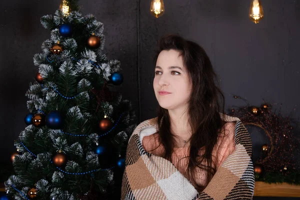 Portrait of attractive brunette woman in beige knitted sweater near christmas tree in dark room. new year theme. cosy holidays and celebration — стоковое фото