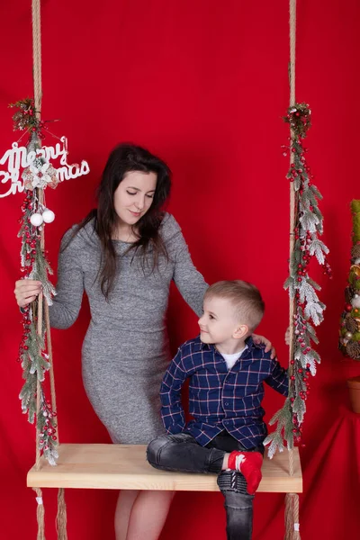 Glad ofullständig familj kramas på trä gunga dekorerad med julgran grenar på röd bakgrund. mysig ung familj. ensamstående mor i grå klänning och liten son — Stockfoto