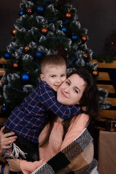 Young mother in a beige knitted sweater near a Christmas tree with her son. single mother with child. family time. mothers love — Stock Fotó