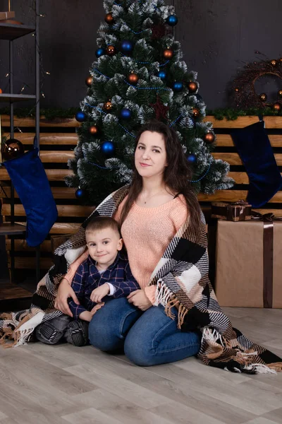 Young mother in a beige knitted sweater near a Christmas tree with her son. single mother with child. family time. mothers love — Stockfoto