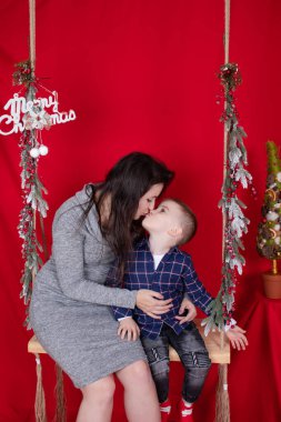 Mutlu eksik aile kucaklaşması ahşap salıncakta kırmızı arka planda noel ağacı dallarıyla süslenmiş. Sıcak, genç bir aile. Gri elbiseli bekar anne ve küçük oğlu.