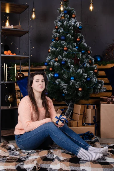 Брюнетка в бежевом трикотажном свитере позирует возле рождественской елки и украшает лофт-комнату голубыми цветами. женский портрет нового года — стоковое фото