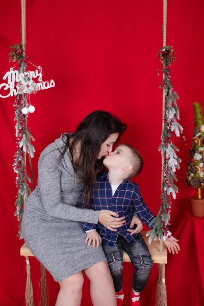 Boldog befejezetlen család ölelés fa hinta díszített karácsonyfa ágak piros háttér. hangulatos fiatal család. egyedülálló anya szürke ruhában és kisbaba fia — Stock Fotó