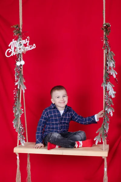Гарненька блондинка сидить на дерев'яній гойдалці, прикрашена гілками дерев і різдвяним декором. на червоному тлі. новорічна тема — стокове фото