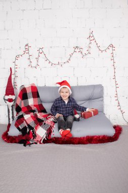 preschool boy in santa hat standing near tall christmas gnome toy, holding little dwarf toy in hand. in grey room. happy christmas and new year. christmas decor and toy