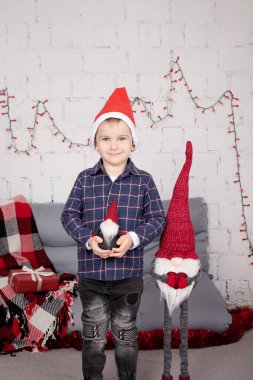 little boy in santa hat near christmas gnome toy, holding dwarf toy in hand in grey room. christmas decor and toys. kids playing