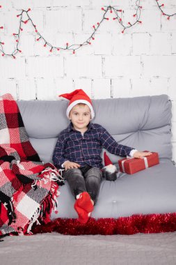 little boy in santa hat near christmas gnome toy, holding dwarf toy in hand in grey room. christmas decor and toys. kids playing