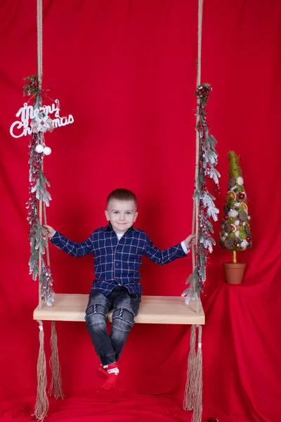 Menino loiro bonito sentado em um balanço de madeira decorado com galhos de árvore e decoração de Natal. no fundo vermelho. tema de ano novo — Fotografia de Stock