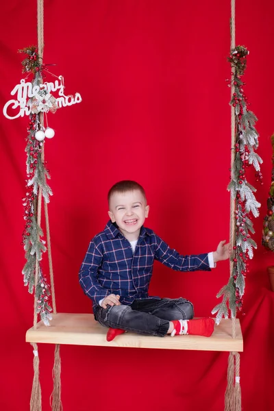 Menino loiro bonito sentado em um balanço de madeira decorado com galhos de árvore e decoração de Natal. no fundo vermelho. tema de ano novo — Fotografia de Stock