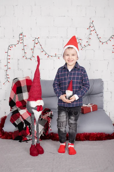 Little boy in santa hat near christmas gnome toy, holding dwarf toy in hand in grey room. christmas decor and toys. kids playing — Zdjęcie stockowe
