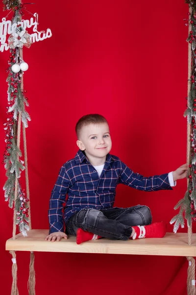 Pretty blonde boy sitting on a wooden swing decorated with tree branches and christmas decor. on red background. new year theme — Stockfoto