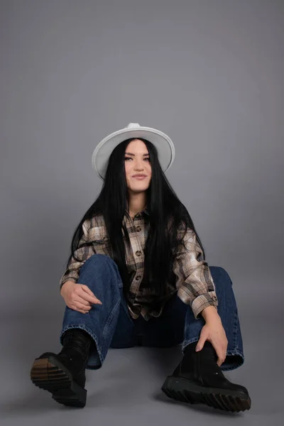 Jovem atraente em camisa xadrez, chapéu branco e jeans em fundo cinza. Retrato feminino bonito. — Fotografia de Stock