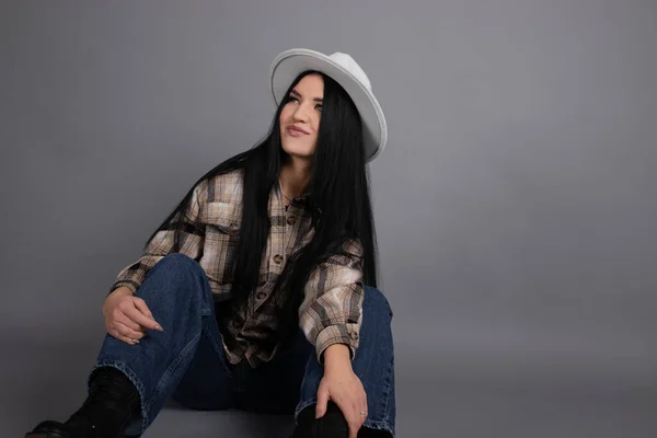 Attractive young woman in plaid shirt, white hat and jeans on gray background. pretty female portrait. — Stock Photo, Image
