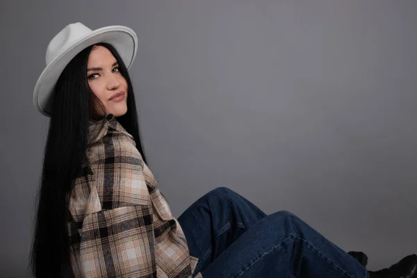 Attractive young woman in plaid shirt, white hat and jeans on gray background. pretty female portrait. — стоковое фото