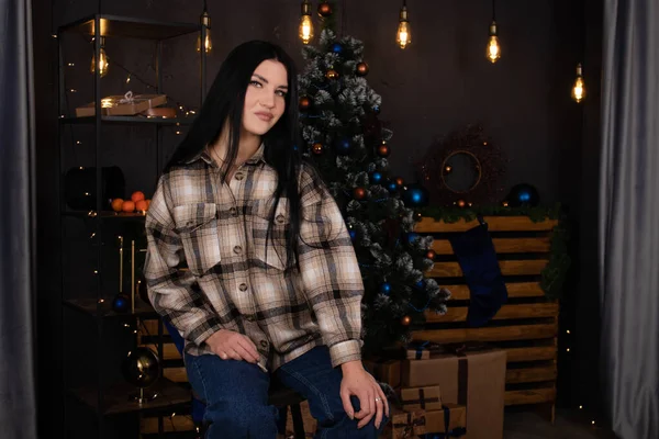 Уверенная красивая молодая женщина в клетчатой рубашке и джинсах возле рождественской елки, украшенной голубыми тонами — стоковое фото