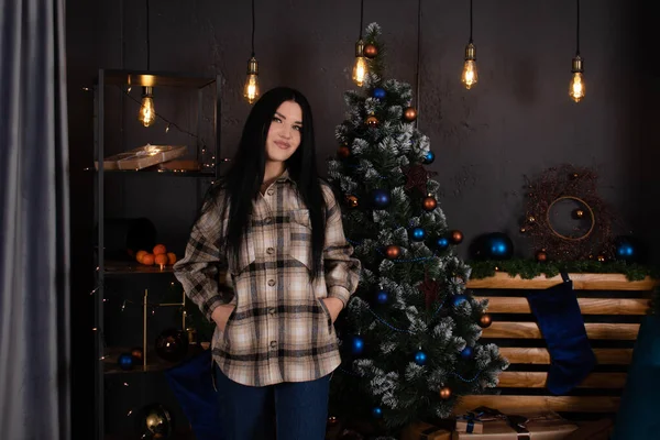 Confident beautiful young woman in checkered shirt and jeans near christmas tree decorated in blue tones — стоковое фото