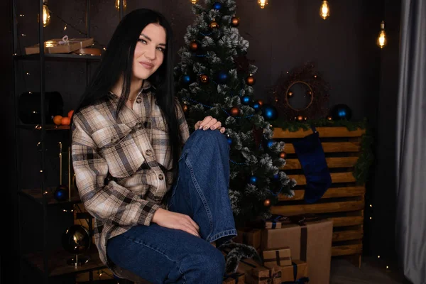 Уверенная в себе красивая молодая женщина в клетчатой рубашке возле рождественской елки. Новый год, зимняя тема — стоковое фото