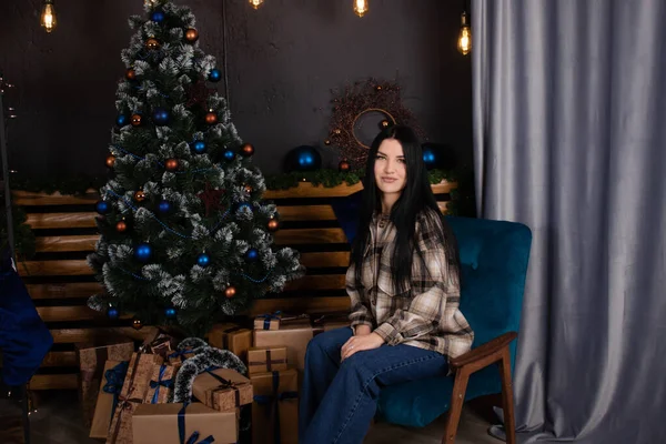 Confident beautiful young woman in checkered shirt and jeans near christmas tree decorated in blue tones — стоковое фото