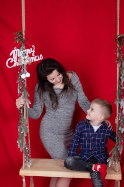 Mutlu eksik aile kucaklaşması ahşap salıncakta kırmızı arka planda noel ağacı dallarıyla süslenmiş. Sıcak, genç bir aile. Gri elbiseli bekar anne ve küçük oğlu.