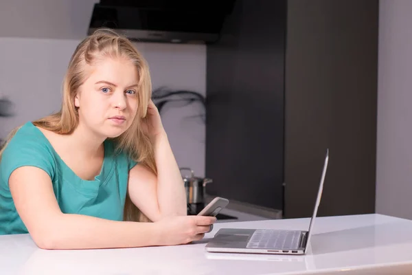 Bonita chica adolescente rubia en camiseta verde trabajando o estudiando cerca de la computadora portátil en habitación gris —  Fotos de Stock