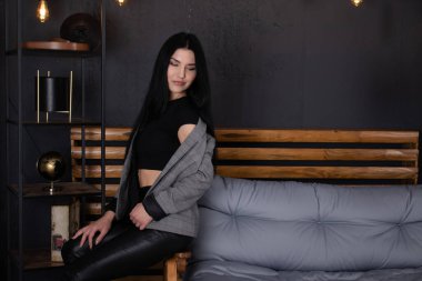 Gri çatı katında, siyah elbiseli, kendine güvenen esmer kadın bir kanepede oturuyor. Moda kıyafetler. moda kadın portresi