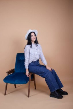 Bluzlu çekici esmer kadın, kahverengi arka planda mavi sandalyede şapka ve kot pantolon. kadın moda portresi