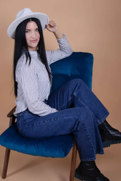 Porträtt av ung kvinna i vit blus, vit hatt och jeans nära blå stol på brun bakgrund. vacker kvinnlig porträtt — Stockfoto
