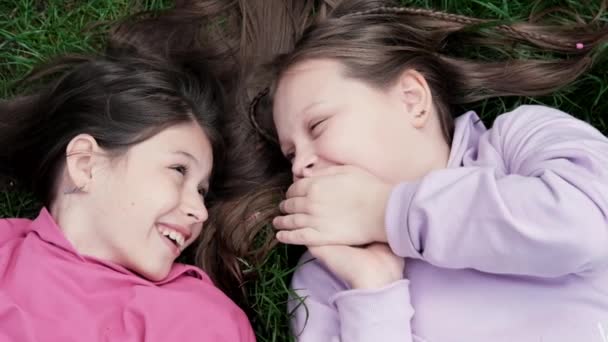 Dos hermosos amigos de los niños del milenio que yacen en la hierba verde en el parque o el césped. Chicas caucásicas sonriendo y divirtiéndose juntos. desde la vista superior — Vídeos de Stock