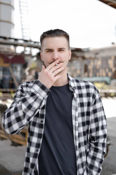 Joven hombre guapo con bigote en camisa a cuadros blanco y negro fumando un cigarrillo fuera. mal hábito —  Fotos de Stock