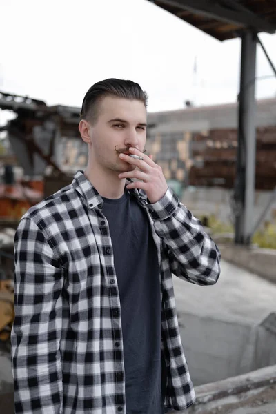 Joven hombre guapo con bigote en camisa a cuadros blanco y negro fumando un cigarrillo fuera. mal hábito —  Fotos de Stock