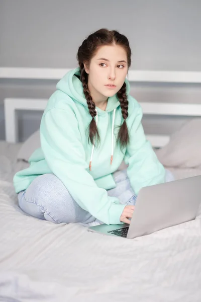 Joven morena con dos coletas en sudadera con capucha turquesa y jeans sentados en una cama con sábanas blancas con portátil. trabajar o estudiar desde casa. restricciones de cuarentena. trabajo lejano, estudio remoto —  Fotos de Stock