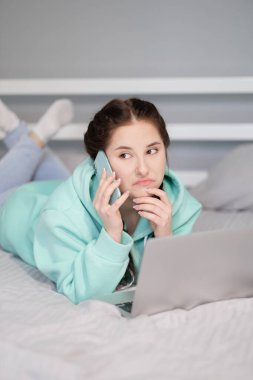 Çevrimiçi eğitim ve çalışma mesafesi. Ofiste çalışan mutlu esmer kadın evden turkuaz kapüşonlu ve yatakta kot pantolonla çalışıyor. Konuşan telefon. Bilgisayar laptopu kullanılıyor.