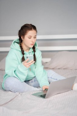 Çevrimiçi eğitim ve çalışma mesafesi. Ofiste çalışan mutlu esmer kadın evden turkuaz kapüşonlu ve yatakta kot pantolonla çalışıyor. Konuşan telefon. Bilgisayar laptopu kullanılıyor.