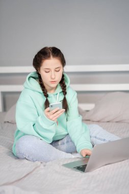 Çevrimiçi eğitim ve çalışma mesafesi. Ofiste çalışan mutlu esmer kadın evden turkuaz kapüşonlu ve yatakta kot pantolonla çalışıyor. Konuşan telefon. Bilgisayar laptopu kullanılıyor.