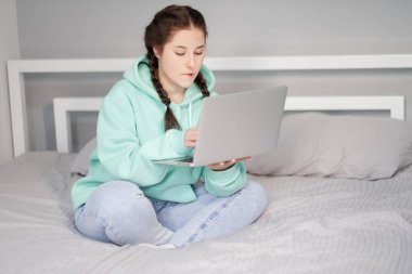 Çevrimiçi eğitim ve çalışma mesafesi. Turkuaz kapüşonlu ve kot pantolonlu mutlu bir kadın evden beyaz yatakta çalışıyor. Bilgisayar laptopu kullanılıyor.