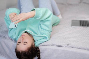 Evde çalışmak ya da ders çalışmak. Turkuaz kapüşonlu genç esmer kadın ve dizüstü bilgisayarı ve turkuaz renkli telefonuyla yatakta kot pantolon. Karantina kısıtlamaları. uzak çalışma, çalışma. Mutlu uzaktan eğitim