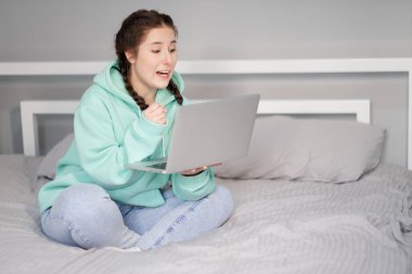 Çevrimiçi eğitim ve çalışma mesafesi. Turkuaz kapüşonlu ve kot pantolonlu mutlu bir kadın evden beyaz yatakta çalışıyor. Bilgisayar laptopu kullanılıyor.