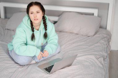 Çevrimiçi eğitim ve çalışma mesafesi. Turkuaz kapüşonlu ve kot pantolonlu mutlu bir kadın evden beyaz yatakta çalışıyor. Bilgisayar laptopu kullanılıyor.