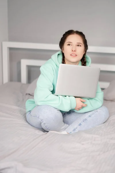 Educación a distancia en línea y trabajo. Mujer feliz chica en sudadera con capucha turquesa y jeans de trabajo de oficina remotamente desde casa en la cama blanca. Uso de ordenador portátil. —  Fotos de Stock