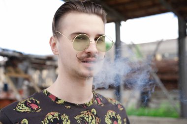 Dışarıda sigara içen bıyıklı, kendine güvenen genç bir adam. Kötü alışkanlık, tütün bağımlılığı