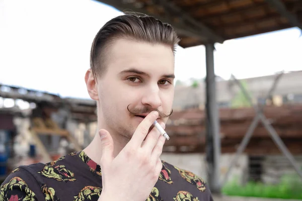 Jovem homem confiante com bigode fumando um cigarro lá fora. mau hábito, vício em tabaco — Fotografia de Stock