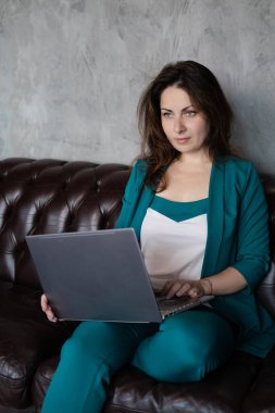 Uzak çalışma, uzaktan eğitim. Deri koltukta dizüstü bilgisayarlı, yeşil elbiseli esmer kadın. Merkez ofis. Evden çalışmak