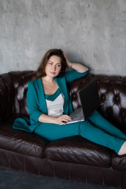 Uzak çalışma, uzaktan çalışma. Yeşil takım elbiseli esmer kadın deri koltukta dizüstü bilgisayarla yalınayak oturuyor. Merkez ofis. Evden çalışmak