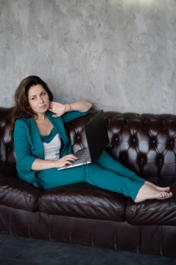 Uzak çalışma, uzaktan çalışma. Yeşil takım elbiseli esmer kadın deri koltukta dizüstü bilgisayarla yalınayak oturuyor. Merkez ofis. Evden çalışmak