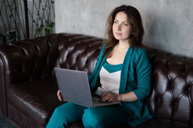Uzak çalışma, uzaktan eğitim. Deri koltukta dizüstü bilgisayarlı, yeşil elbiseli esmer kadın. Merkez ofis. Evden çalışmak
